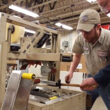 Two men, one in a khaki work shirt and one in a navy work shirt discussing and working on a tape sealing piece of packaging equipment.