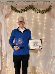 woman in a blue pullover and glasses holding a certificate and plaque, smiling.