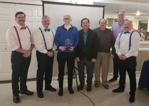 several folks dressed up for an awards banquet surrounding a woman in blue holding a plaque and certificate