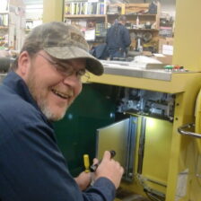 A man wearing a navy work shirt and hat using a flashlight to look into an industrial strapping machine.